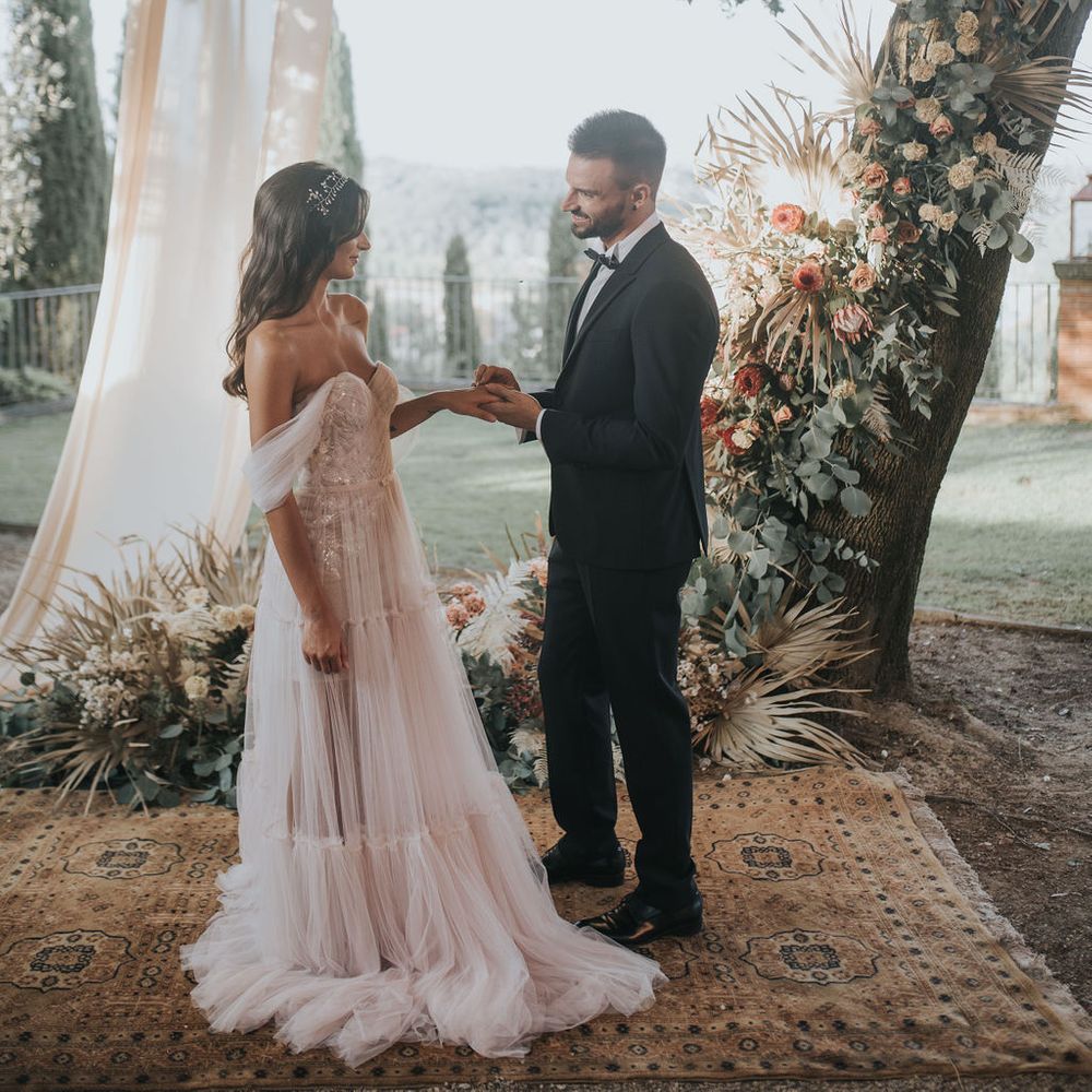 Groom in tuxedo holding his brides hand in front of a tree draped in fabric and decorated with flowers 