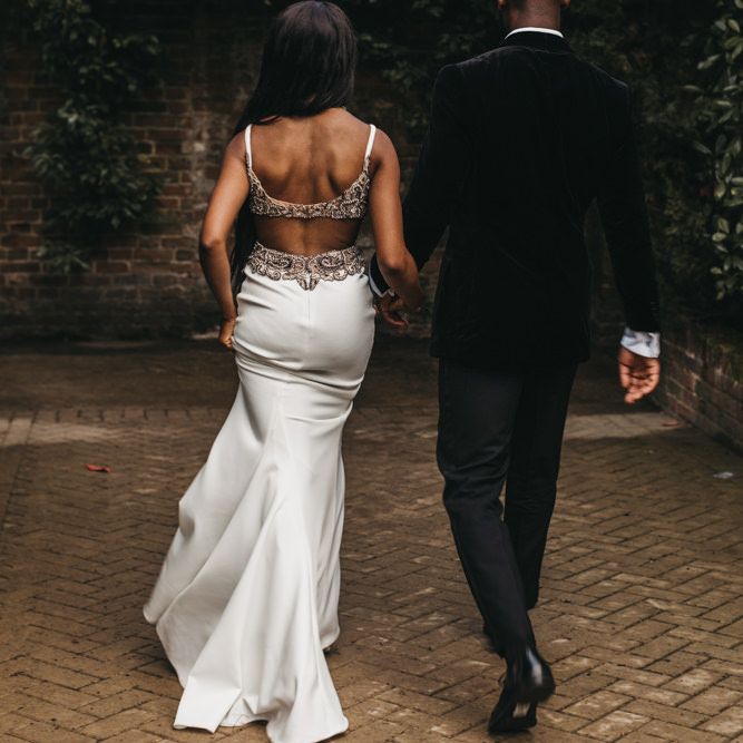 Black bride in a fitted wedding dress with low back detail and embellished straps holding her husbands hand in a velvet jacket 