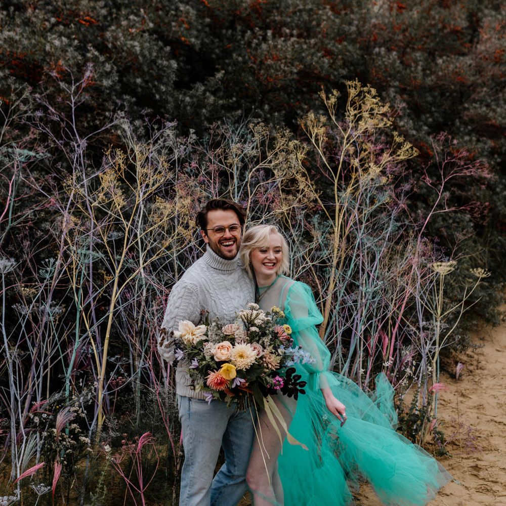 Barefoot bride and groom at their beach elopement standing in the sand by a colourful flower wall holding a bright wedding bouquet 