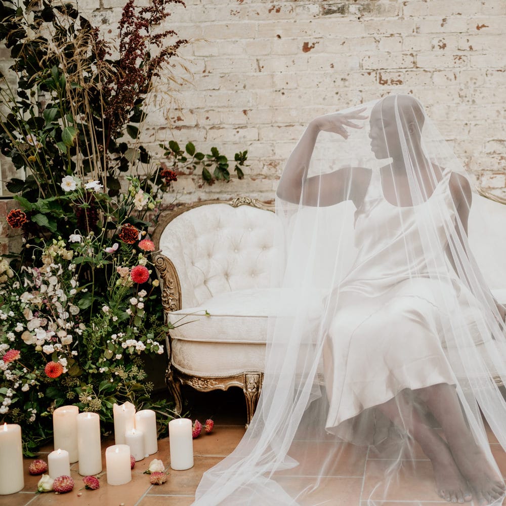 Black bride with alopecia sitting on an ornate sofa in Our Beautiful Glasshouse wearing a satin wedding dress and sheer wedding veil 