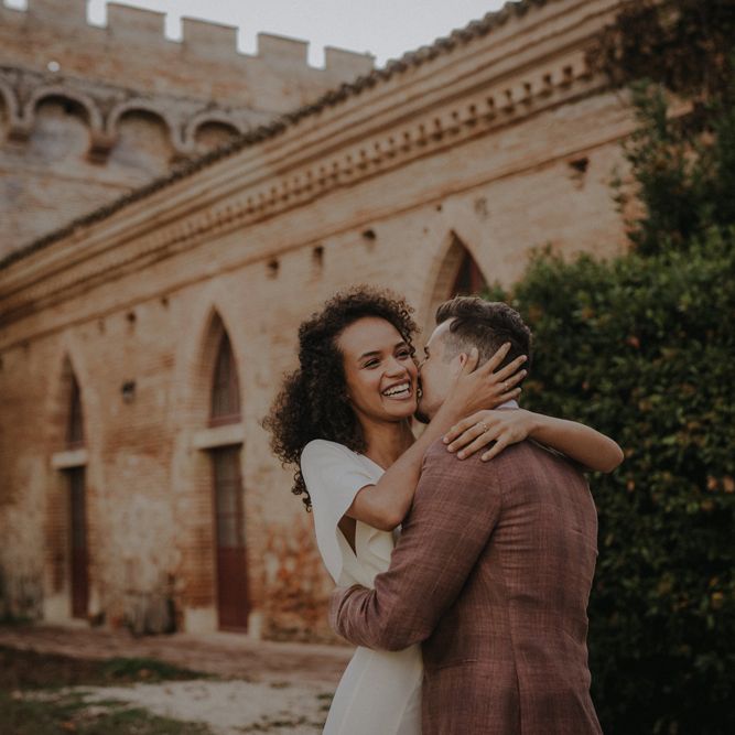 Groom in a brown check suit kissing his smiley bride with naturally curly hair 
