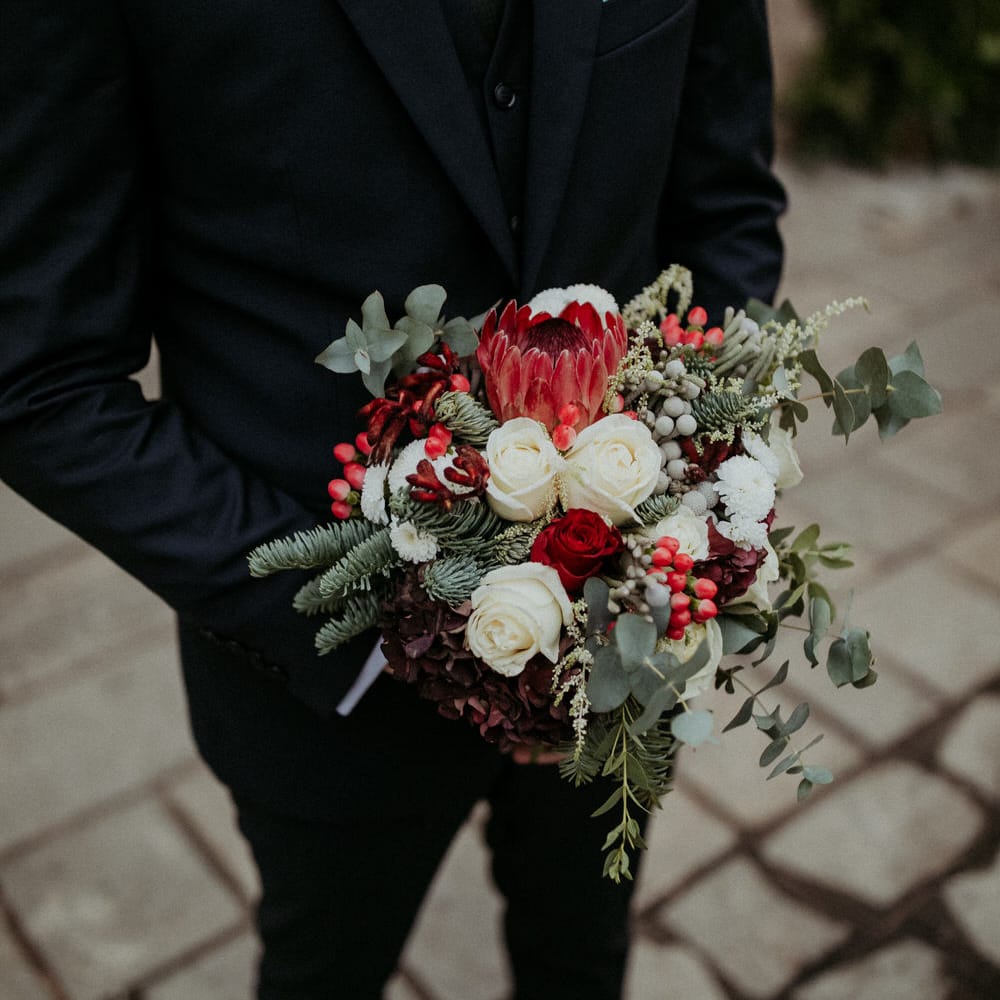 The groom holding a bouquet of red and white roses, eucalyptus and pine leaves, berries and seasonal blooms
