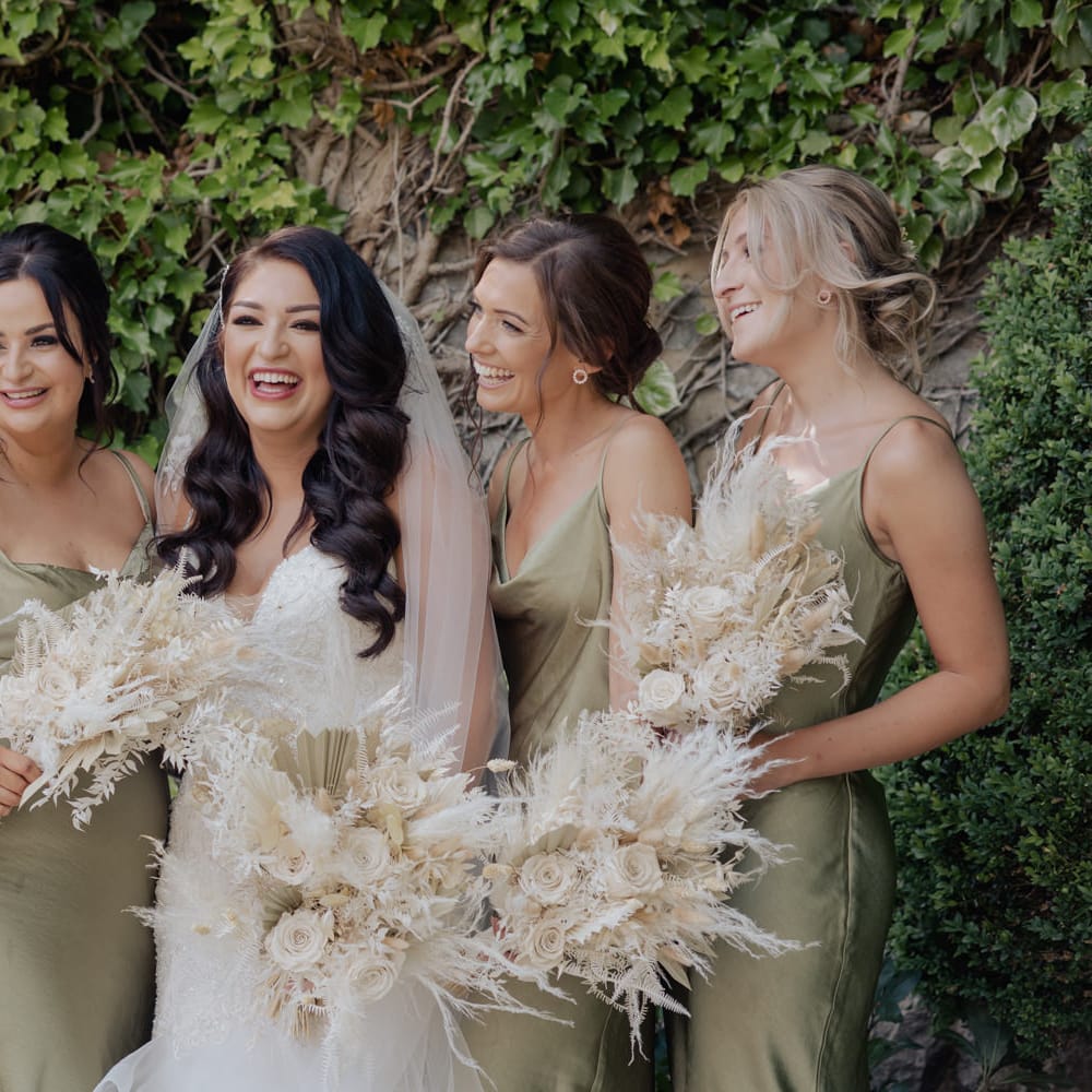 Bridesmaids in sage green satin dresses holding neutral dried flower wedding bouquets standing next to the bride 