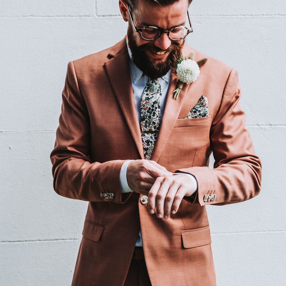 Groom adjusts his shirt sleeve on wedding day as he wears coral coloured suit complete with colourful tie and floral buttonhole 