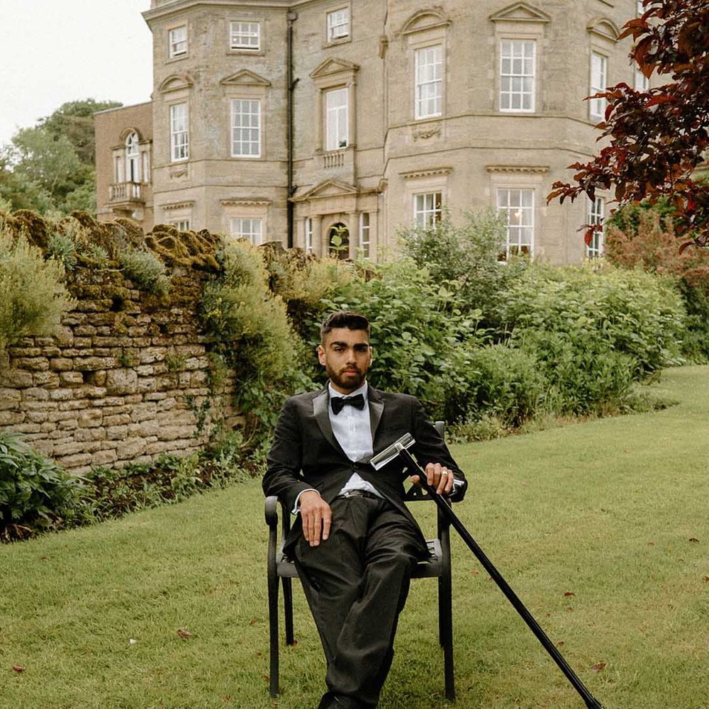 Stylish interabled groom in a black tuxedo sitting on a chair in the gardens at Bourton Hall with his walking stick 