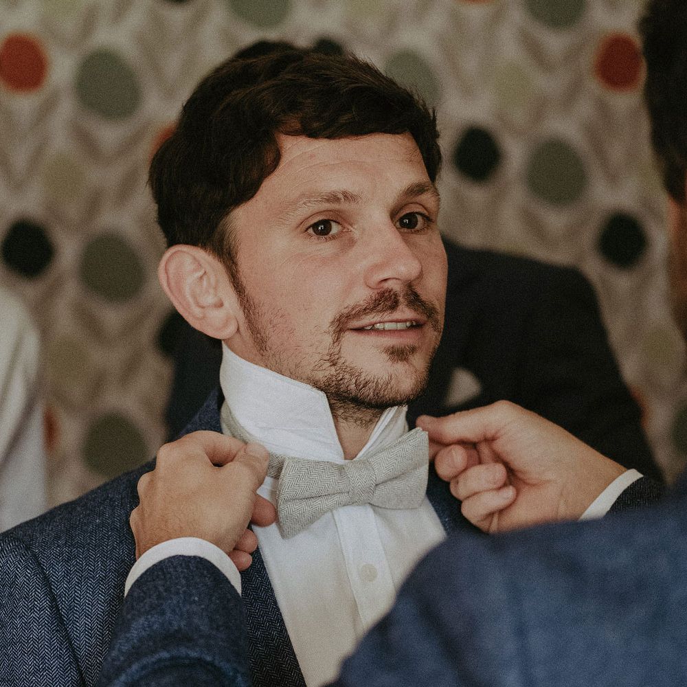 Groom in blue suit jacket and white shirt has his sage green bowtie adjusted by groomsman in blue suit jacket before Isle of Wight wedding with macrame wedding decor