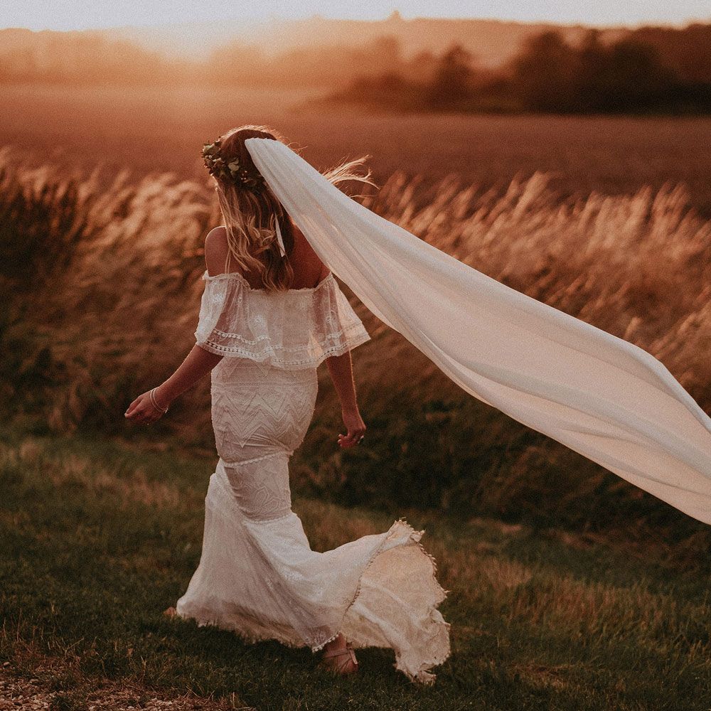 Bride in Grace Loves Lace bardot wedding dress, veil and flower crown walks by field at golden hour with groom in white shirt at Isle of Wight wedding with macrame wedding decor