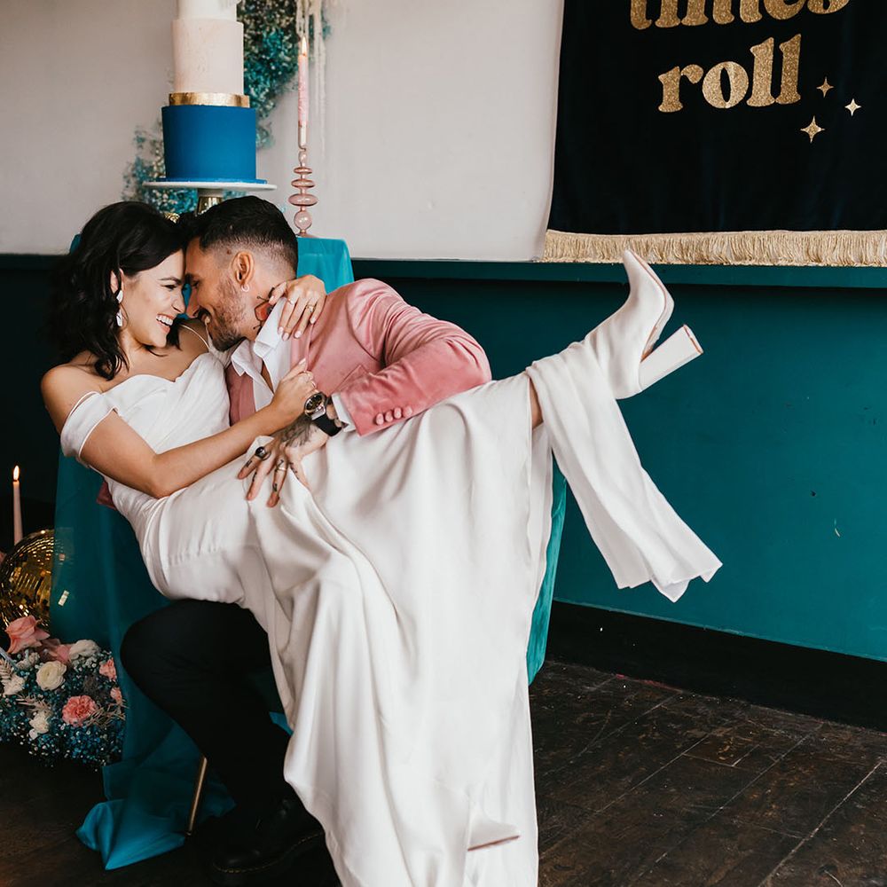 Bride in white wedding boots sitting on her grooms lap at their stylish pink and teal wedding reception 