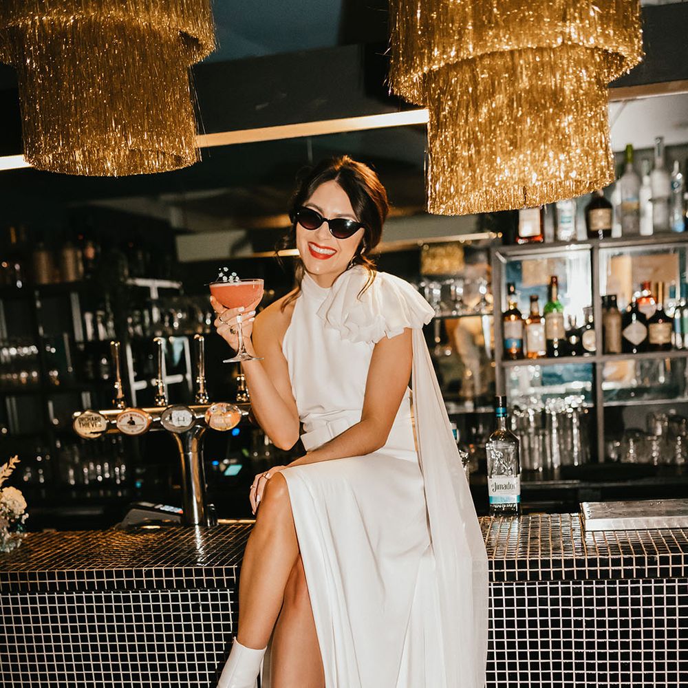 Bride in sunglasses with red lipstick sitting on the bar sipping cocktails in a wedding dress with front slit and white boots 