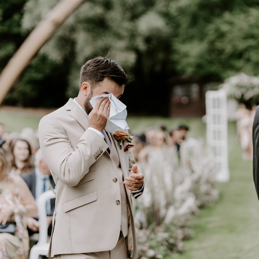 Groom in beige three piece Moss Bros. suit with floral buttonhole wipes eyes with white handkerchief whilst he waits for bride during DIY garden wedding ceremony