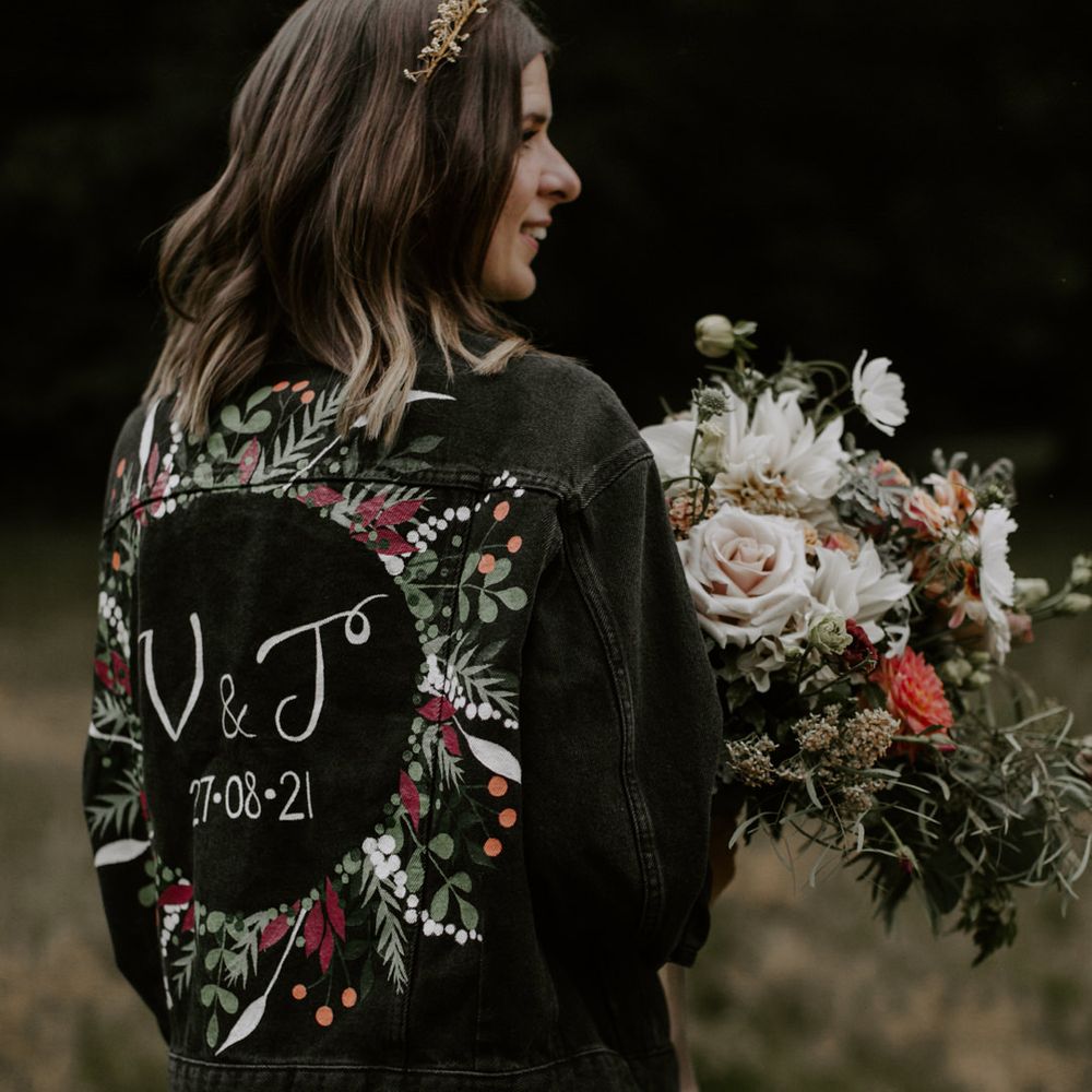 Bride wears personalised denim jacket with floral illustrations whilst holding her bouquet