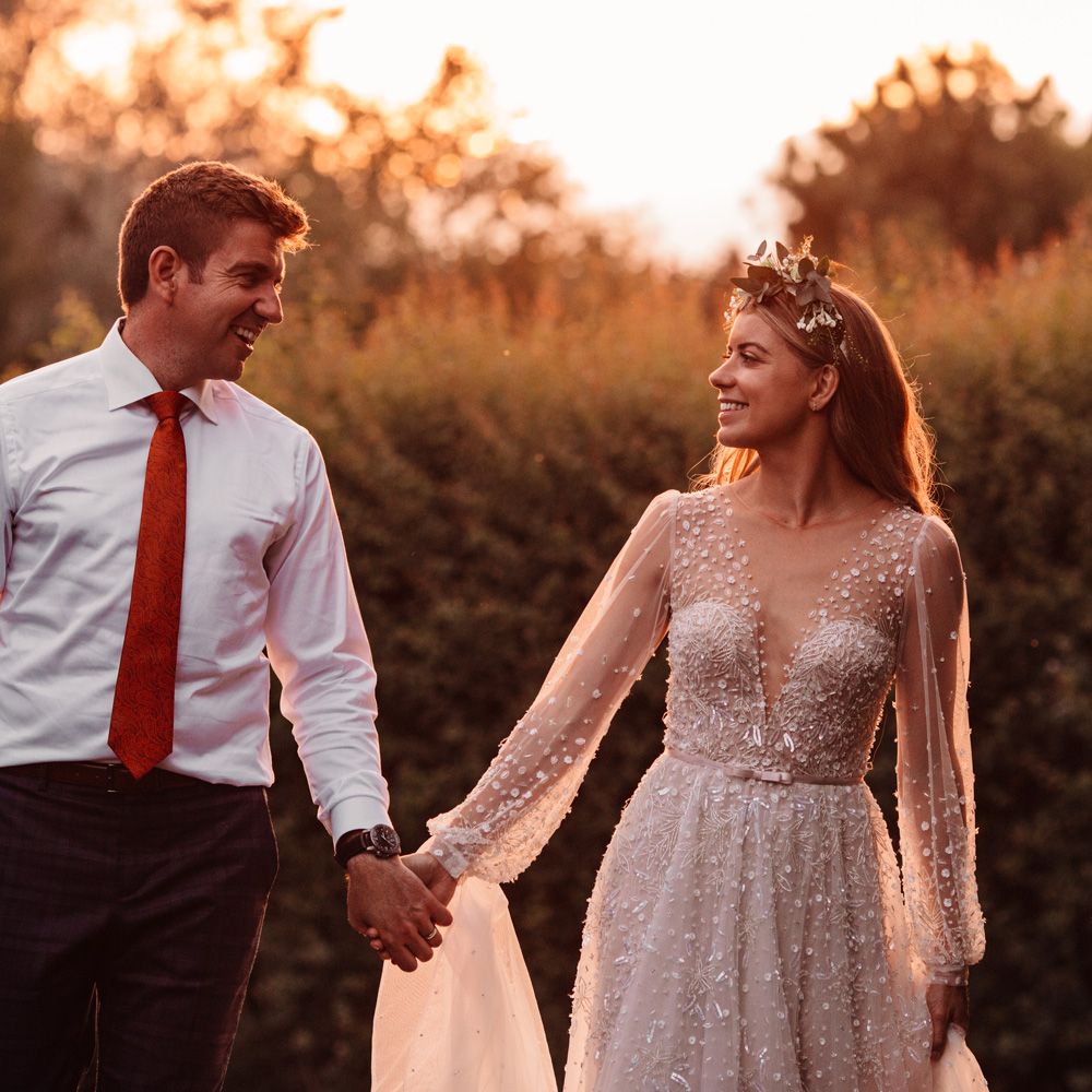 Bride in long mesh sleeve wedding dress with v neck and flower crown smiles whilst holding hands with groom in white shirt and red tie during golden hour