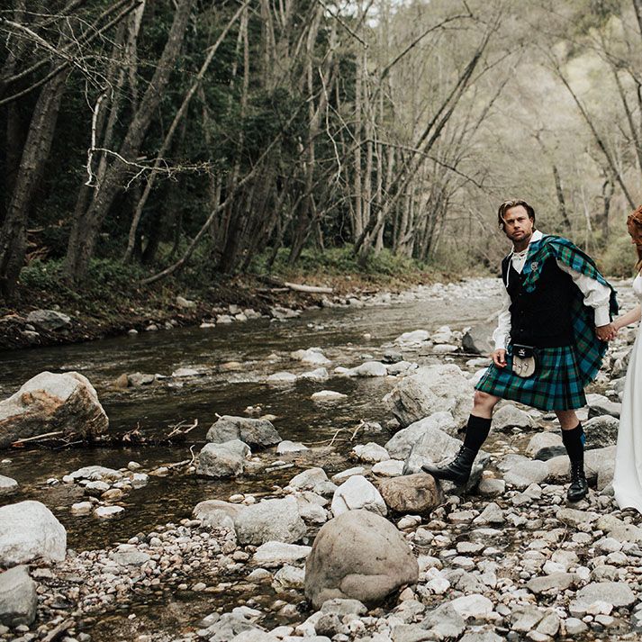 Groom in a tartan kilt and aran jumper holding hands with his bride in an a line wedding dress as they cross a steam at Big Sur California 