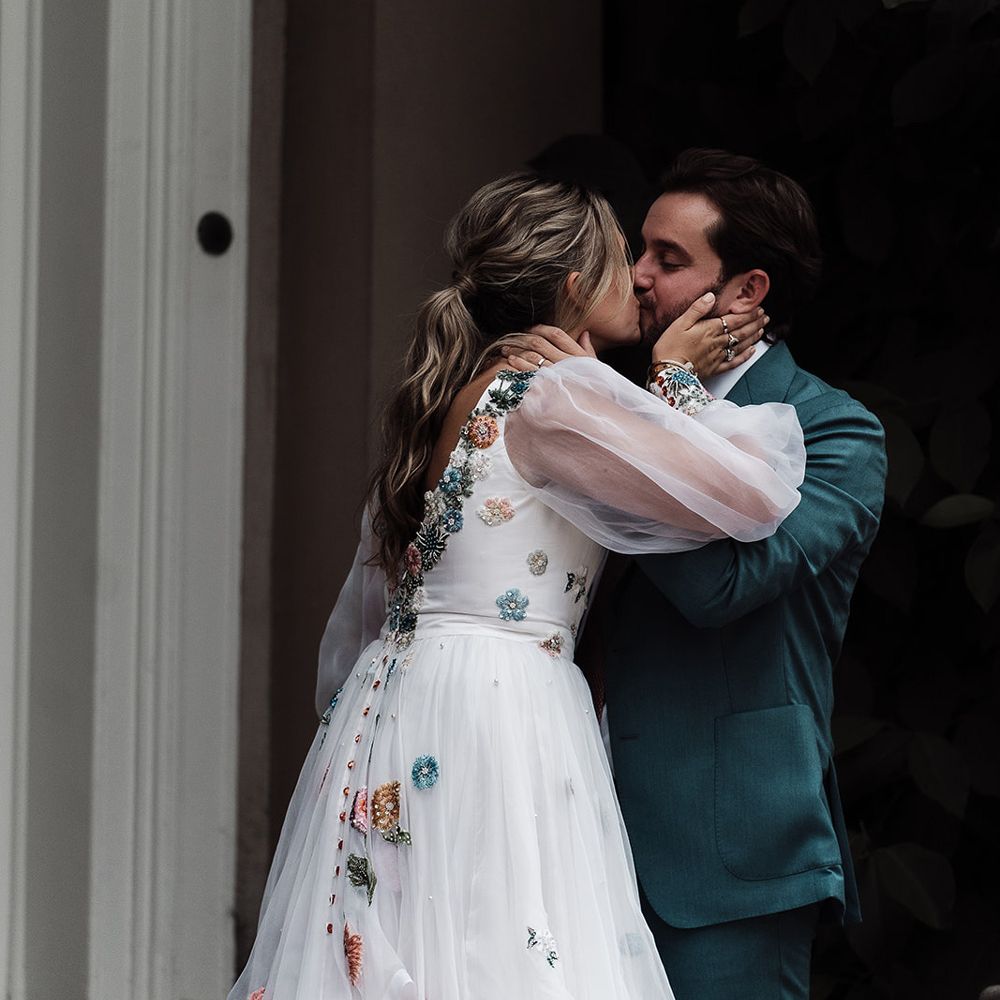 Bride in long mesh sleeve Victoria Sanders wedding dress with low back and floral embroidery kisses groom in teal suit in the grounds of Iscoyd Park
