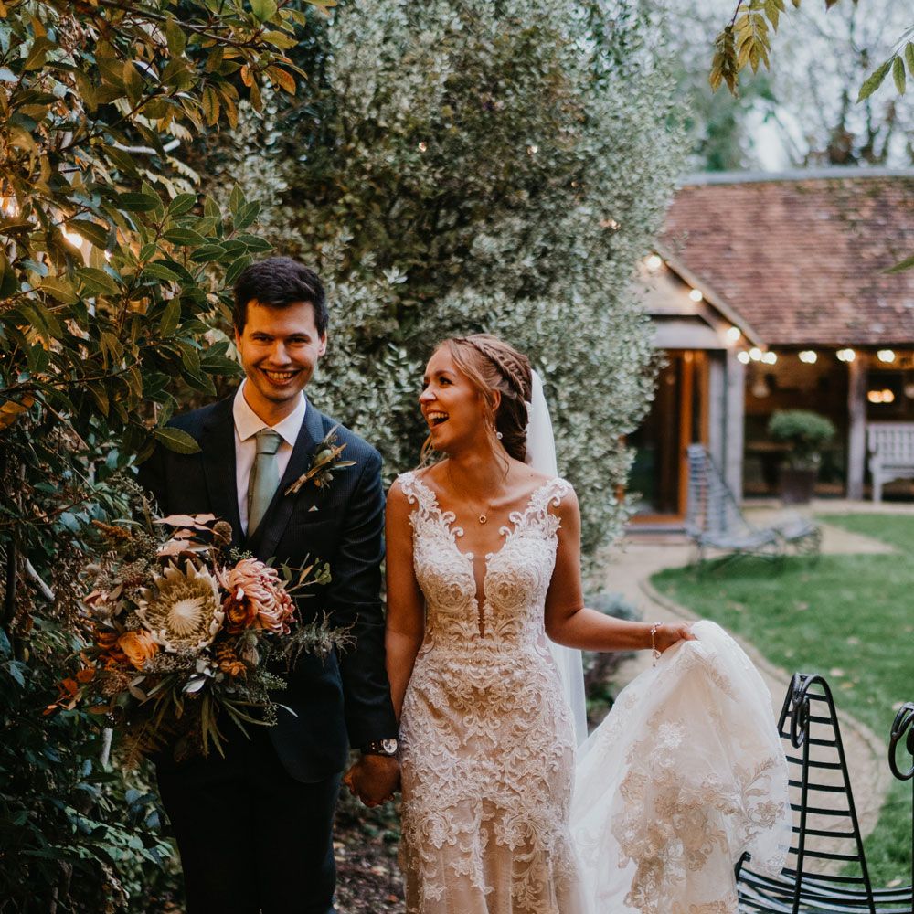 Bride in braided updo and lace wedding dress holding tulle train walks with groom in dark suit and sage green tie holding large bridal bouquet as they walk through gardens