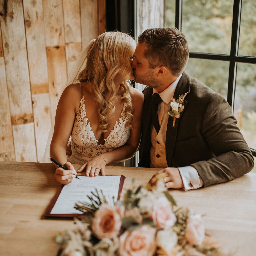 Bride in lace wedding dress and veil sits kissing groom in dark suit with white rose buttonhole as they sign marriage registry at barn wedding