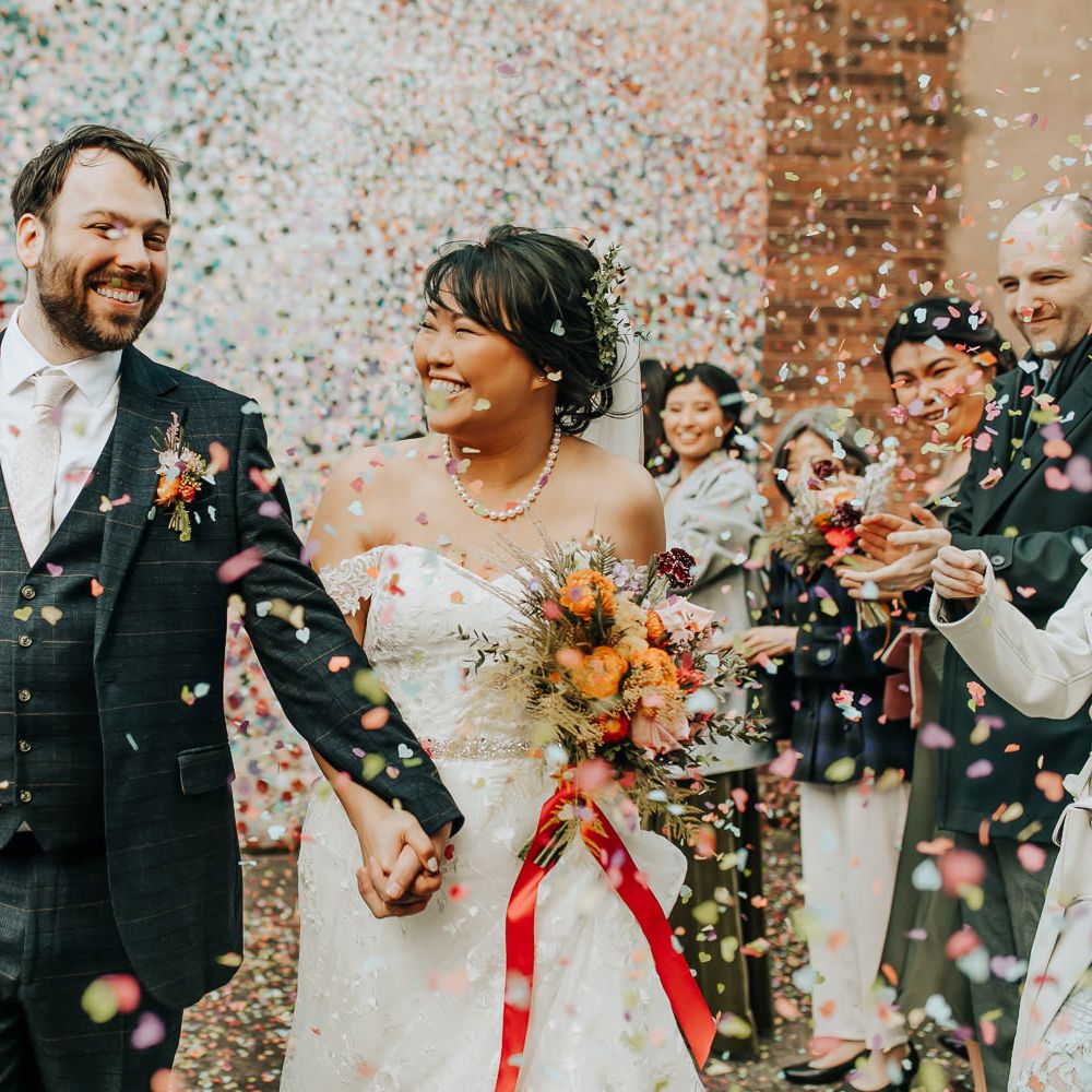 Groom in blue check three piece suit and light pink tie walks holding hands with bride in lace off the shoulder wedding dress and pearl necklace holding mixed bouquet tied with red ribbon as guests throw multi coloured confetti