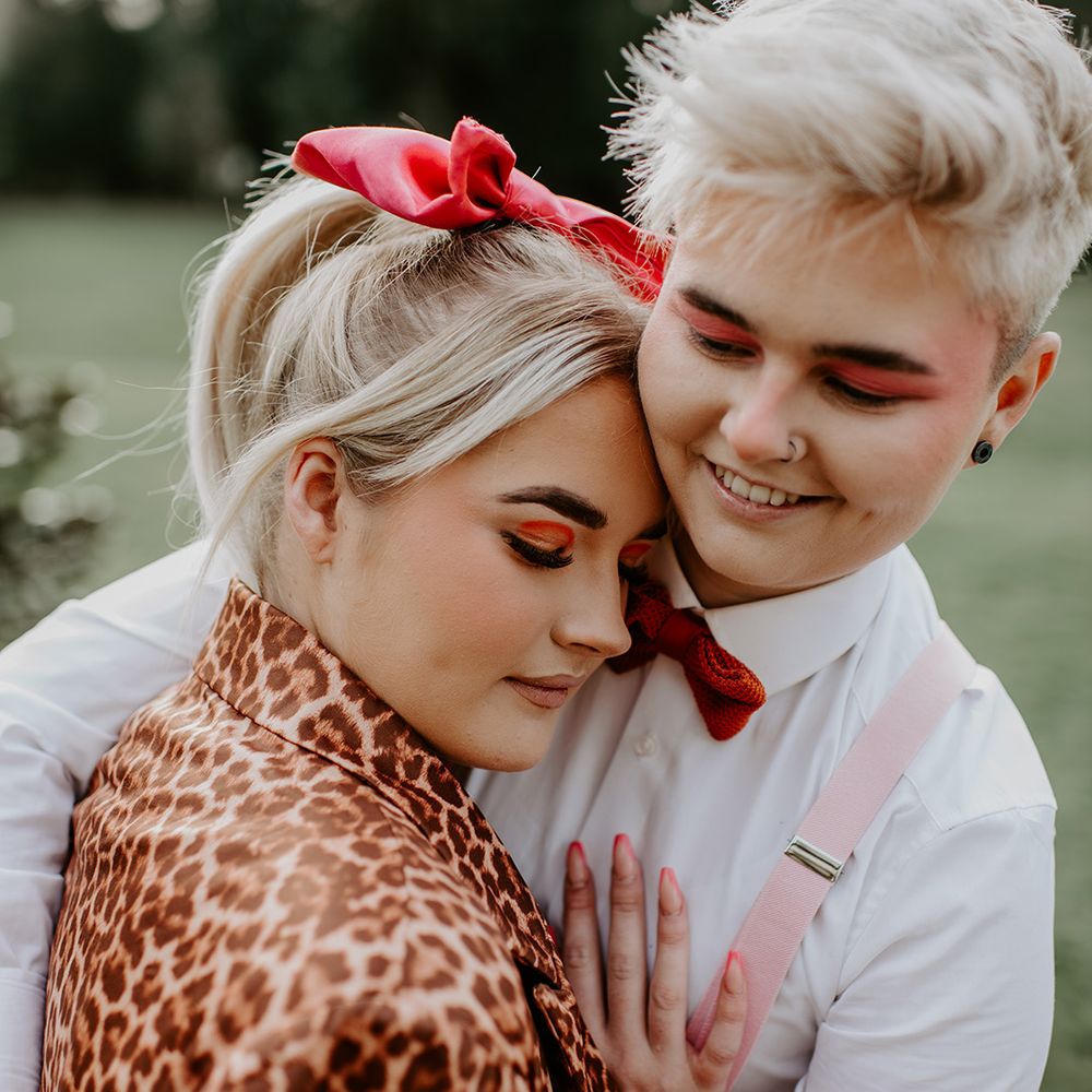 Two brides with red eye makeup embracing with pink braces, red ribbon hair accessory and leopard print jacket 