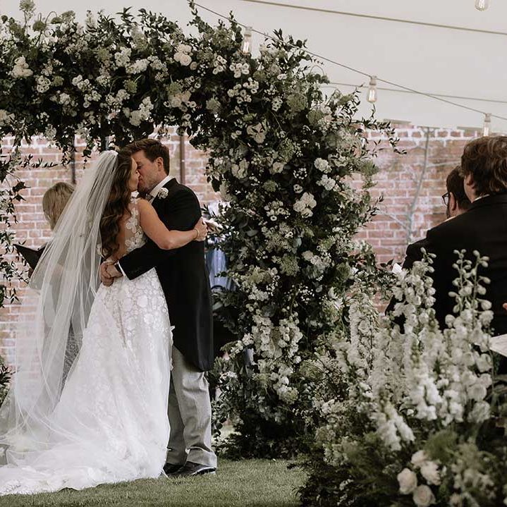 Bride and groom have their first kiss as a married couple in front of large white flower wedding arch for outdoor marquee wedding 