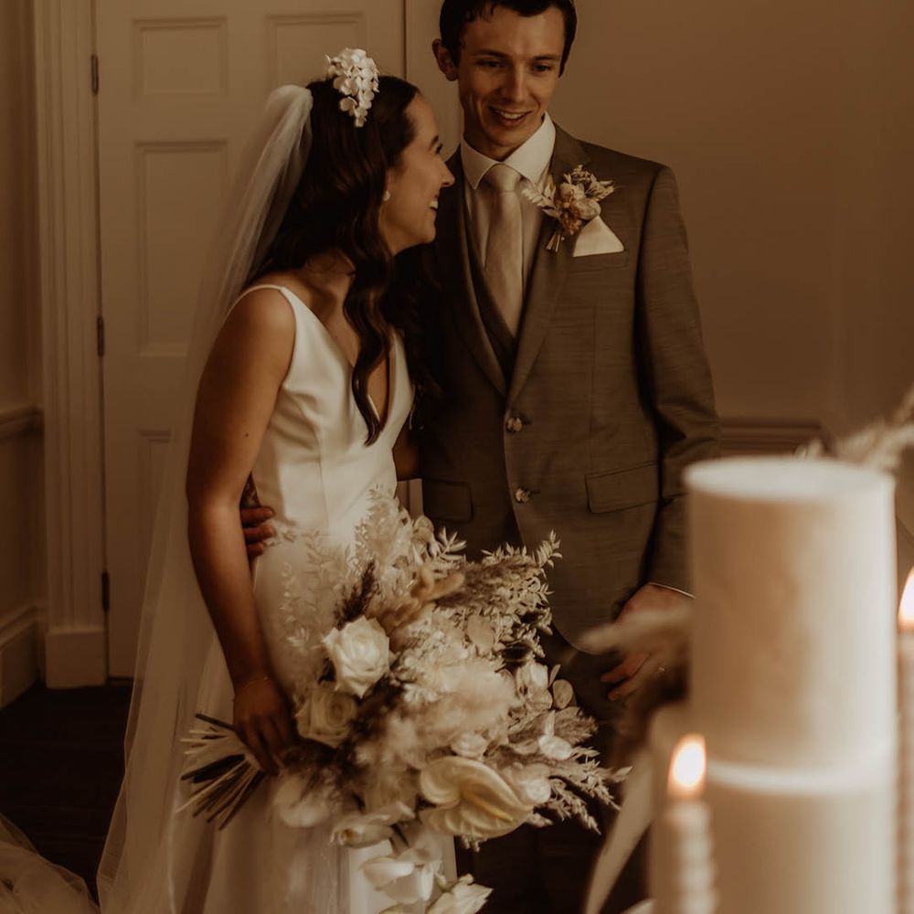 Bride and groom smile and laugh as they take a look at their white wedding cake 