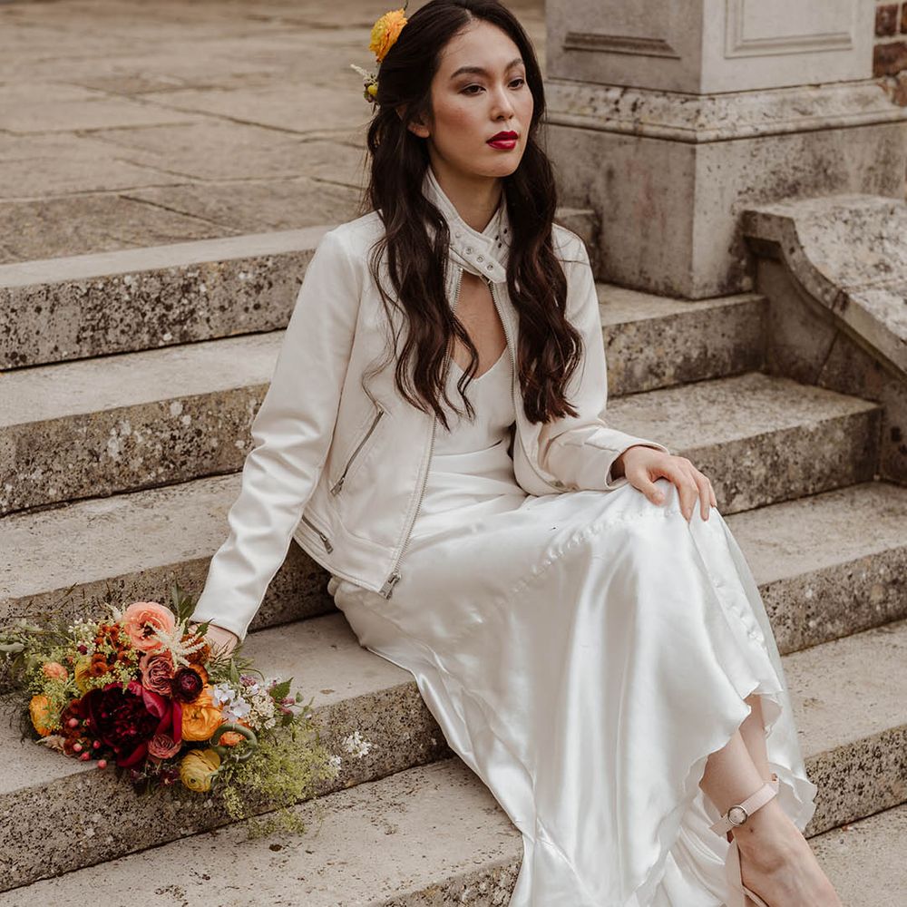 East Asian bride with long wavy hair wearing a flower in har hair, red lipstick, a satin slip wedding dress and a white leather jacket sitting on the steps at Gorse Hill