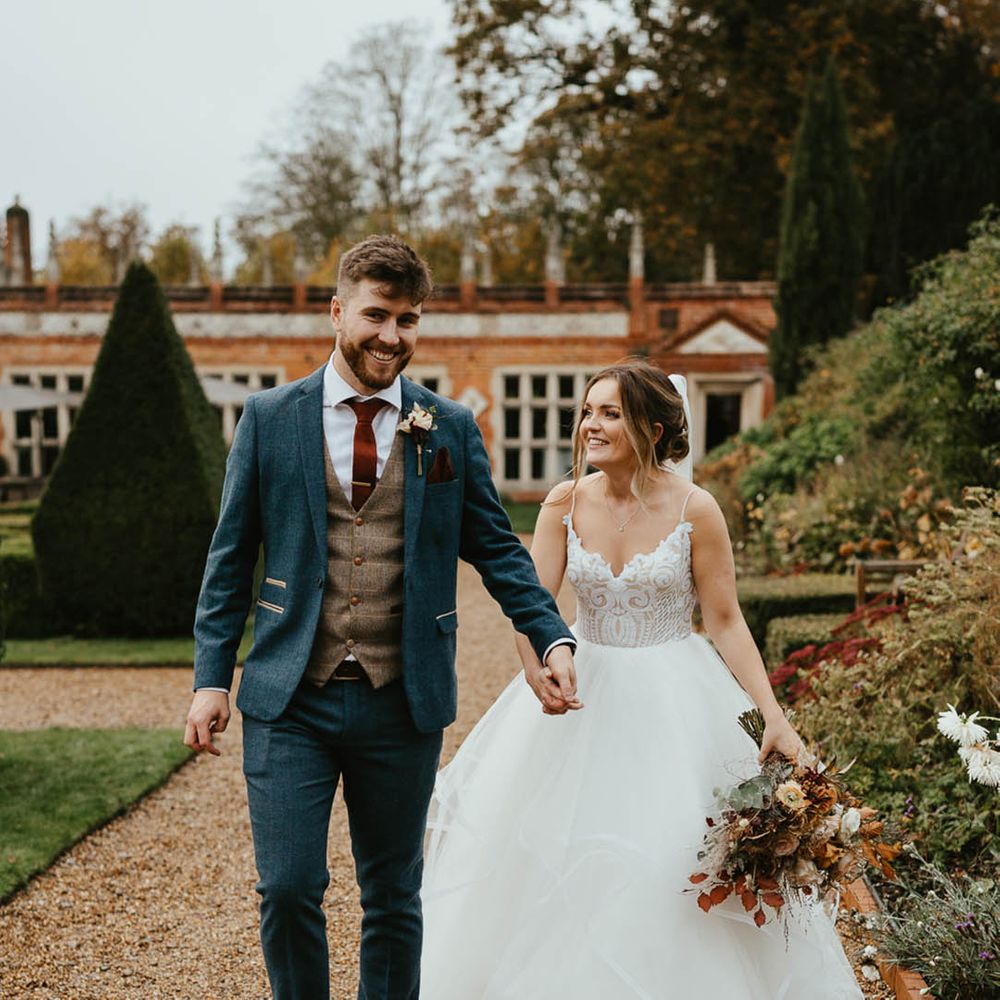 Groom in blue suit with brown waistcoat and burnt orange tie with bride in layered tulle wedding dress walking together around Oxnead Hall 