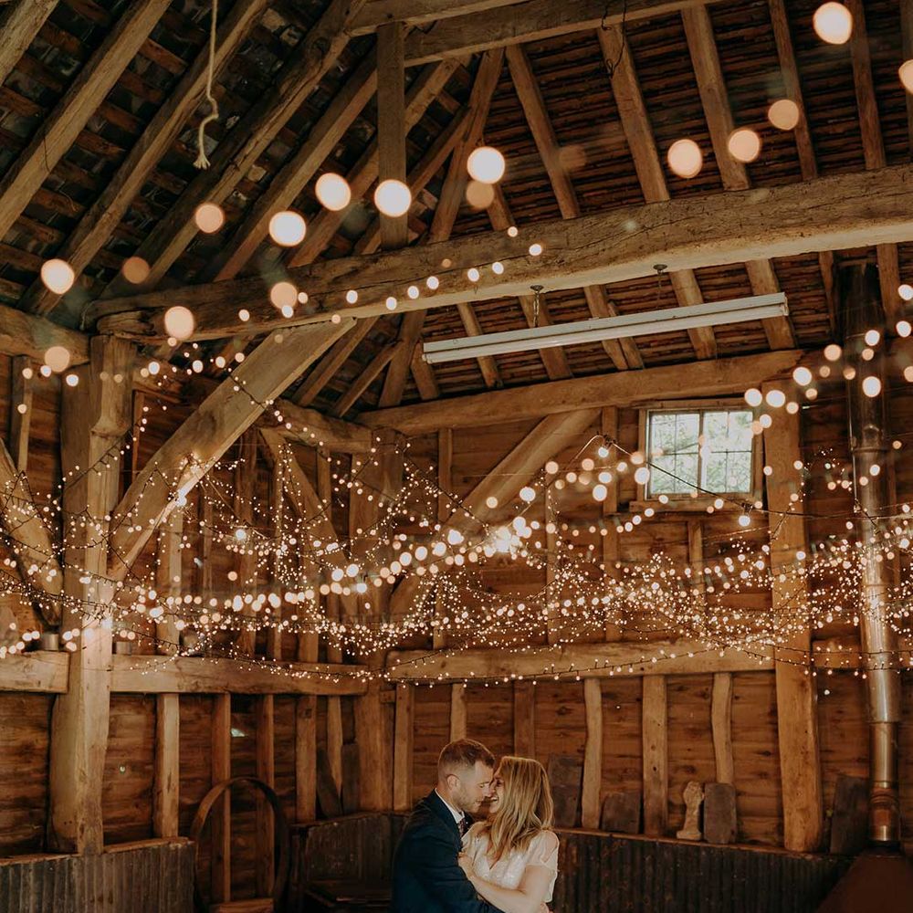 Bride in short sleeved embellished wedding dress and groom in dark suit dancing in a barn covered in fairy lights