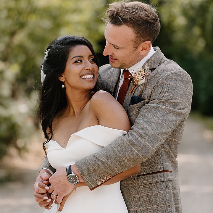Groom in a brown suit hugs the bride from behind in a fitted off the shoulder wedding 