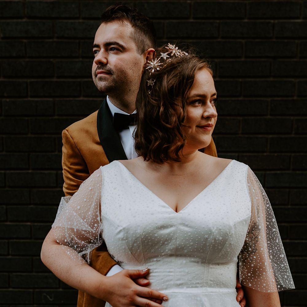 Bride and Groom pose in front of dark brick wall wearing a burnt orange velvet blazer and celestial wedding dress and crown