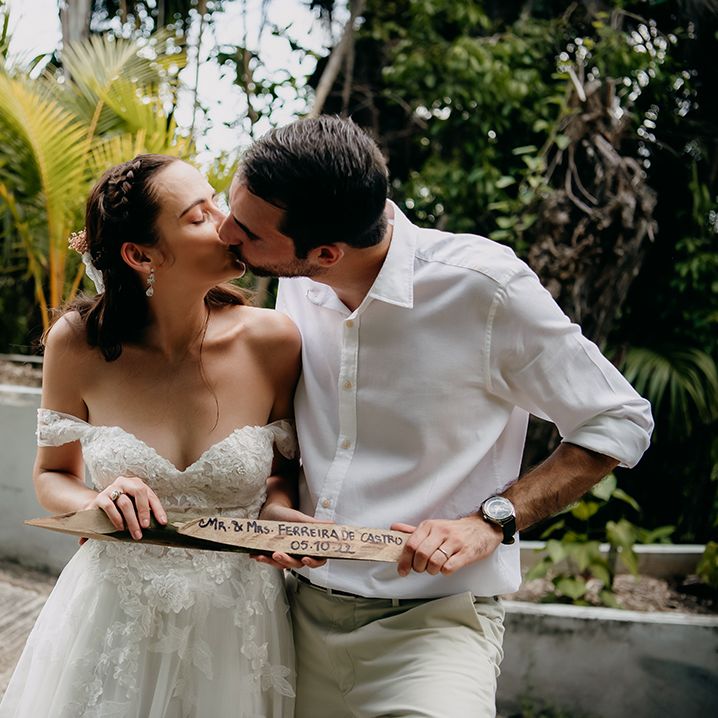 Bride in off the shoulder lace wedding dress with sweetheart neckline and lace floral details and groom in crisp white shirt holding personalised plant stick