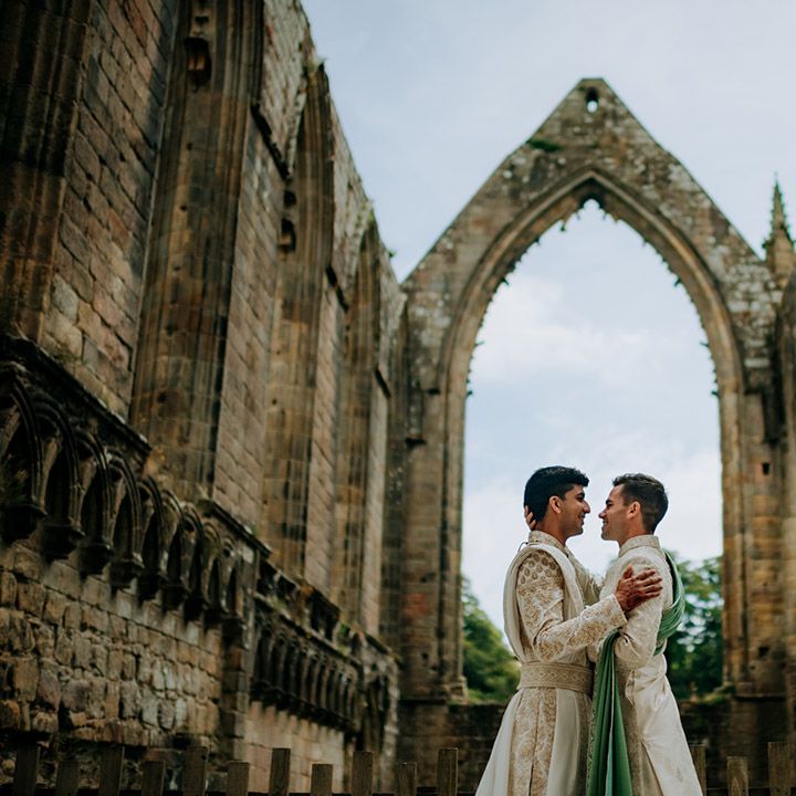 Grooms embrace outdoors during couples portraits surrounded by castle ruins after barn wedding