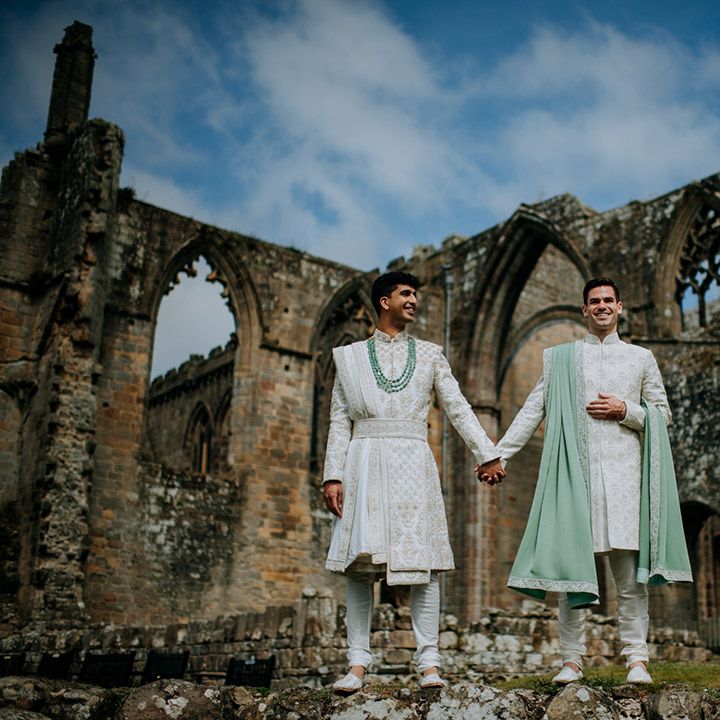 Grooms wear matching sherwani's in white and sage green whilst stood on ruins outdoors during couples portraits