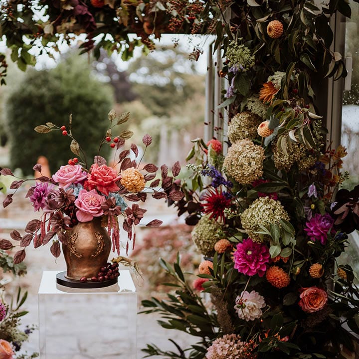 Luxe autumnal flower arch surrounding single tier iced wedding cake with intricate leaf details in the style of an aged golden vase filled with handmade wafer paper florals, pink roses, red roses and yellow carnations 