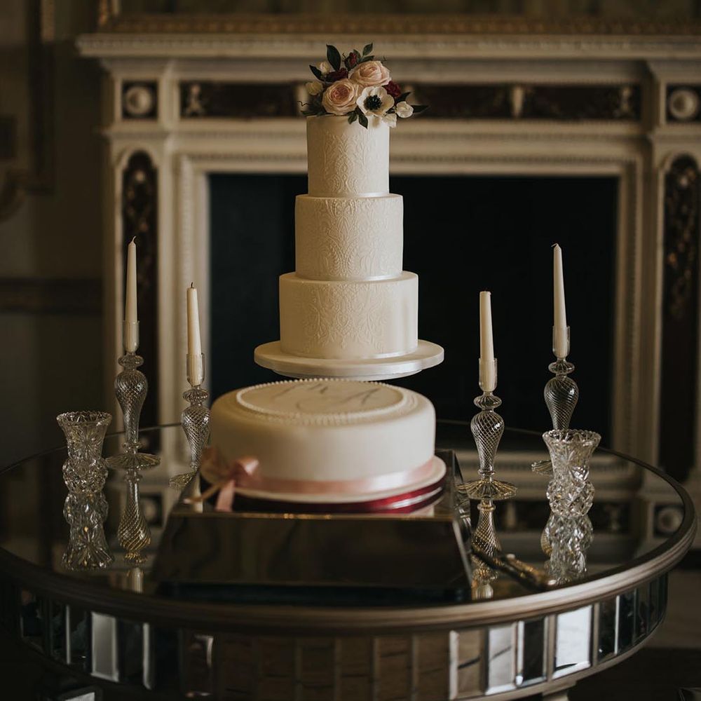 Simple white iced single tier wedding cake with pink ribbon and initials with a larger white iced patterned three tier wedding cake with pink flower decor