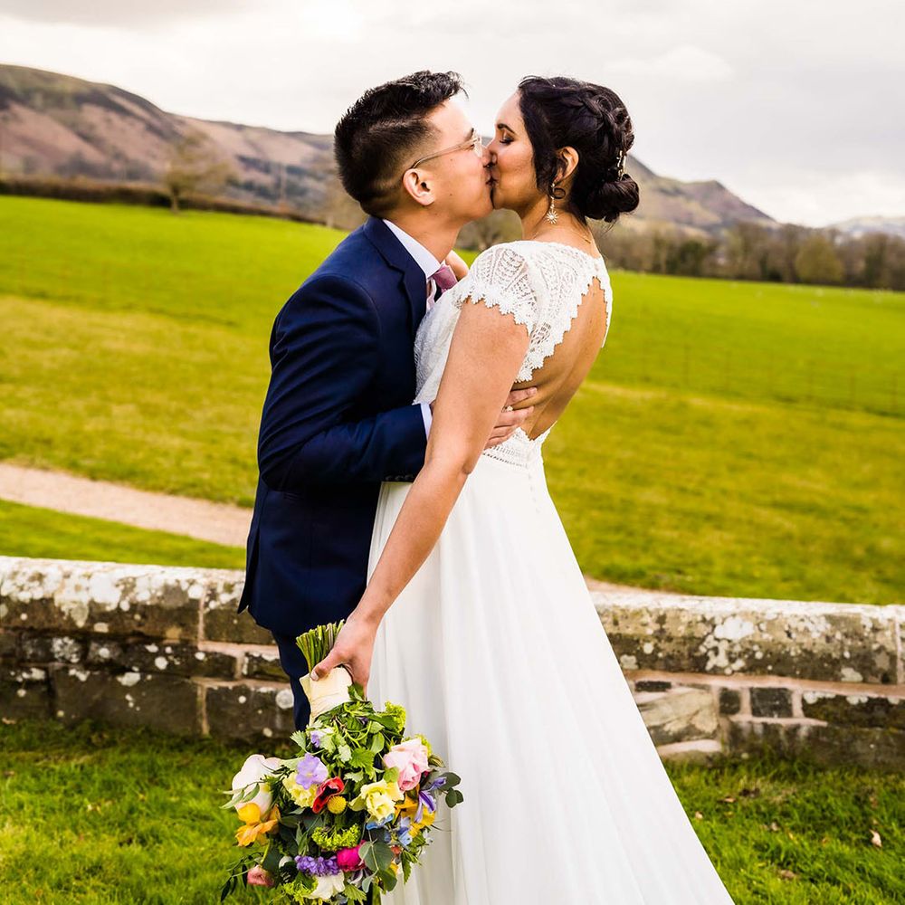 Bride wears lace open back wedding dress whilst holding colourful floral bouquet and kissing her groom outdoors 