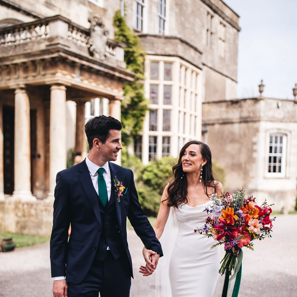 Bride in fitted cowl neck satin Kyha wedding dress with pearl earrings holding colourful bouquet walking with groom in navy suit outside Elmore Court