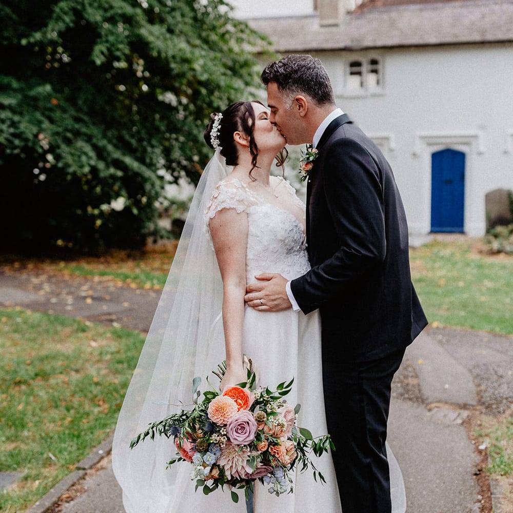 The bride with a flower hairpiece and flower wedding dress holding pastel wedding bouquet kisses the groom in black tie 