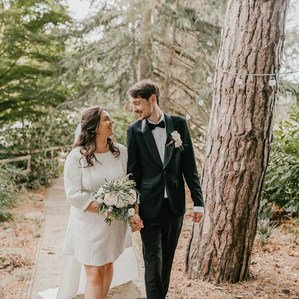 Bride in patterned short wedding dress with train and long sleeves with shoulder length veil walking with the groom in a black tuxedo 