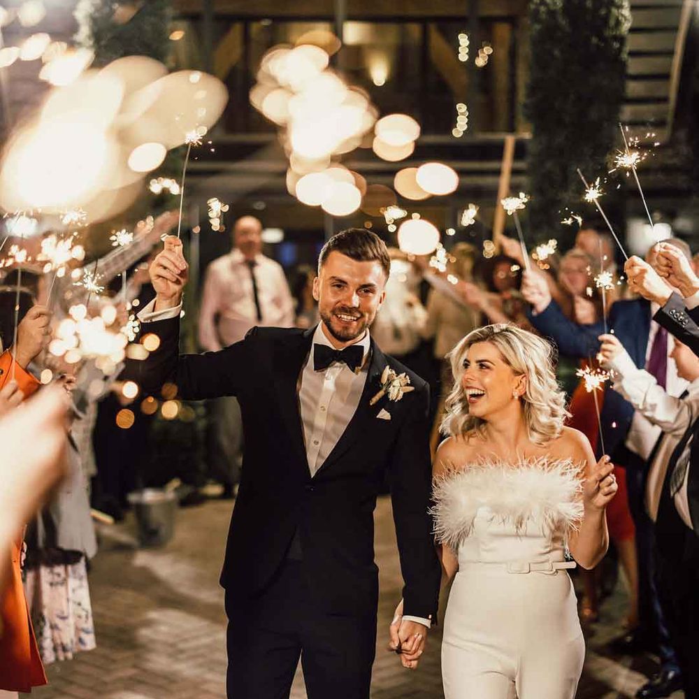 Bride in strapless feather jumpsuit and groom in classic black tux doing sparkler exit at wedding 