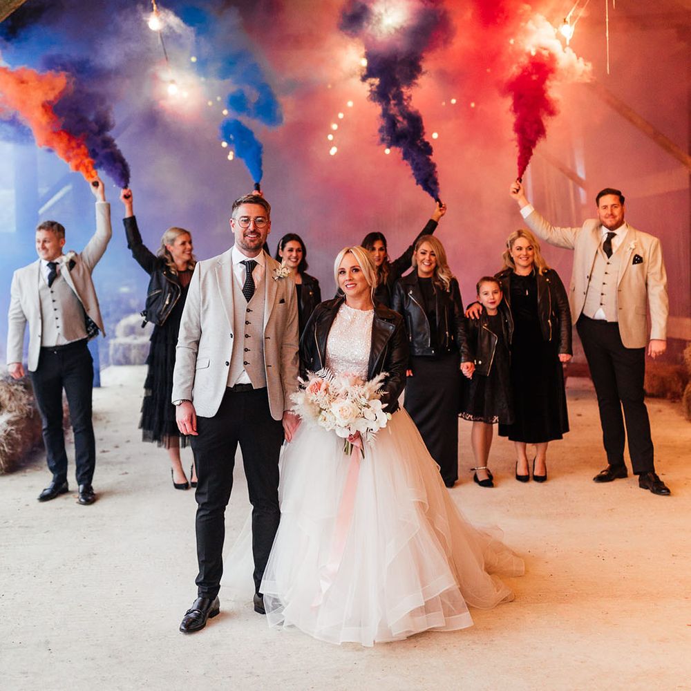Smoke bomb portrait of the bridesmaids in black bridesmaid dresses, groomsmen and groom in matching light greys suits and the bride in sparkly bridal separates