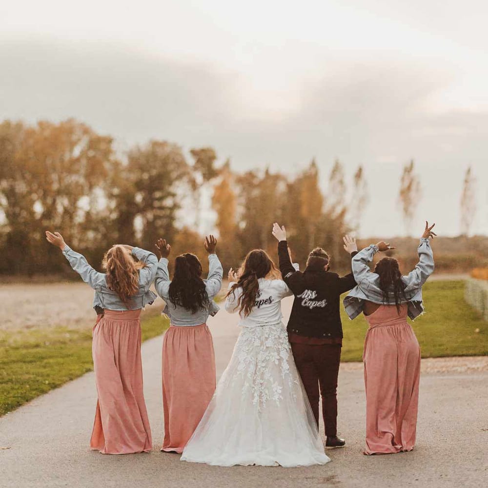 Bridal party wearing personalised denim jackets on top of dusky pink one shoulder bridesmaid dresses and illusion sleeve wedding dress and maroon bridal suit 