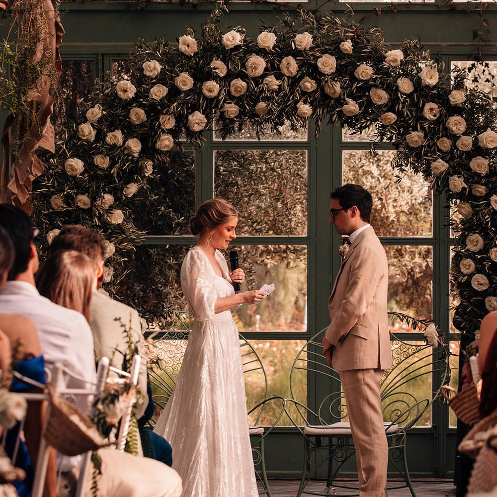 Bride in Grace Loves Lace bridal separates reading out her wedding vows to the groom standing in front of a white rose floral arch 