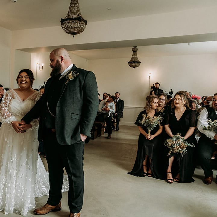 Groom standing at the altar in a green tweed wedding suit with the bride in a long sleeve lace wedding dress 