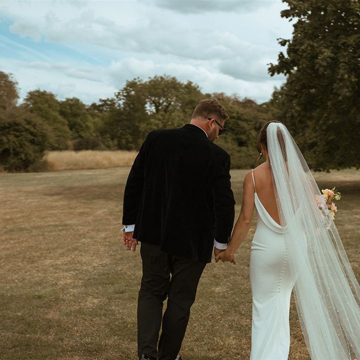 Bride in low back satin wedding dress with button back detail with pearl wedding veil walking along with groom in black tie 