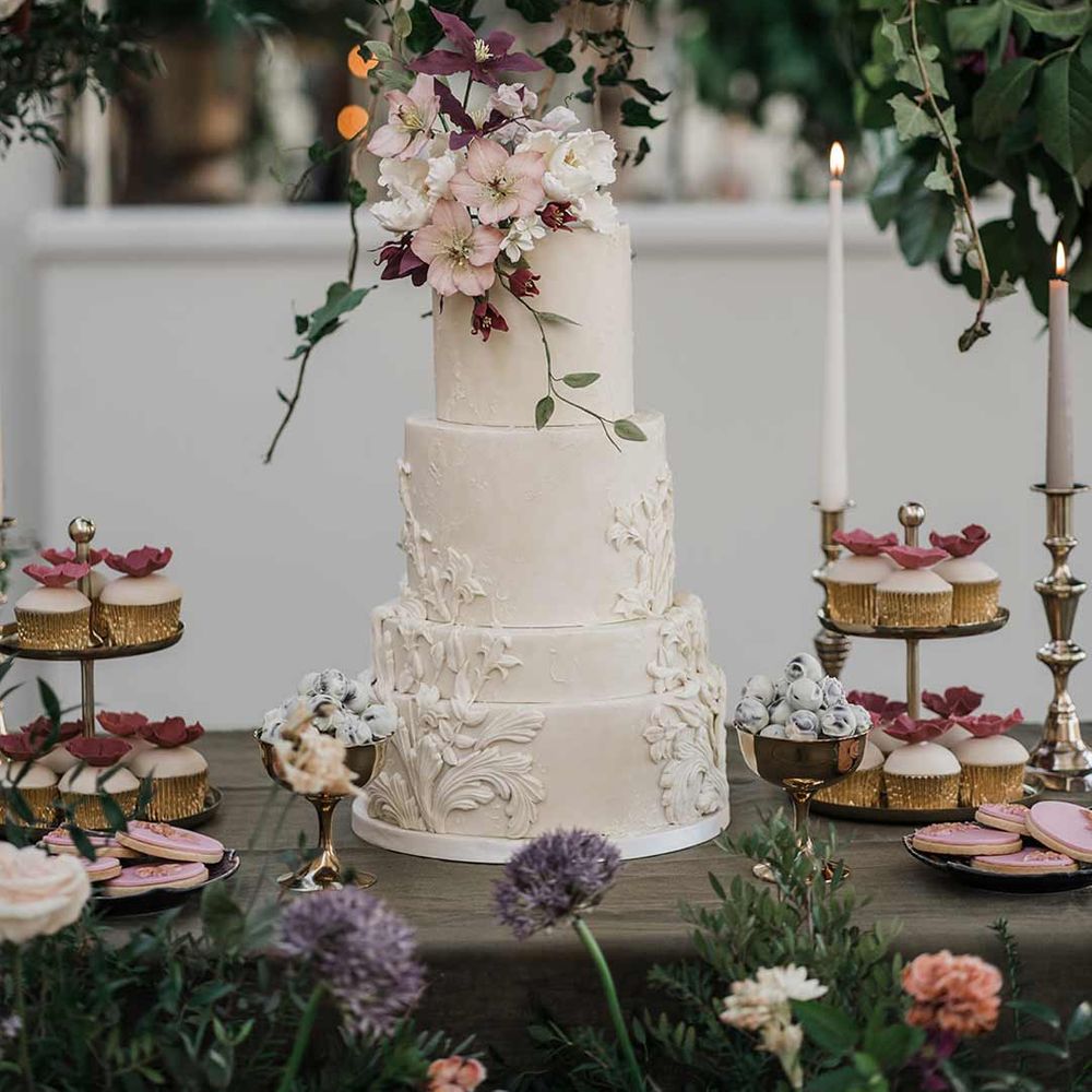 Four tier white wedding cake with intricate icing details and dried flower wedding cake topper on wooden dessert table with wedding cupcakes and wedding biscuits with twisted tapered candles in neutral shades in gold candlesticks surrounded by wildflower and foliage wedding flower arrangements