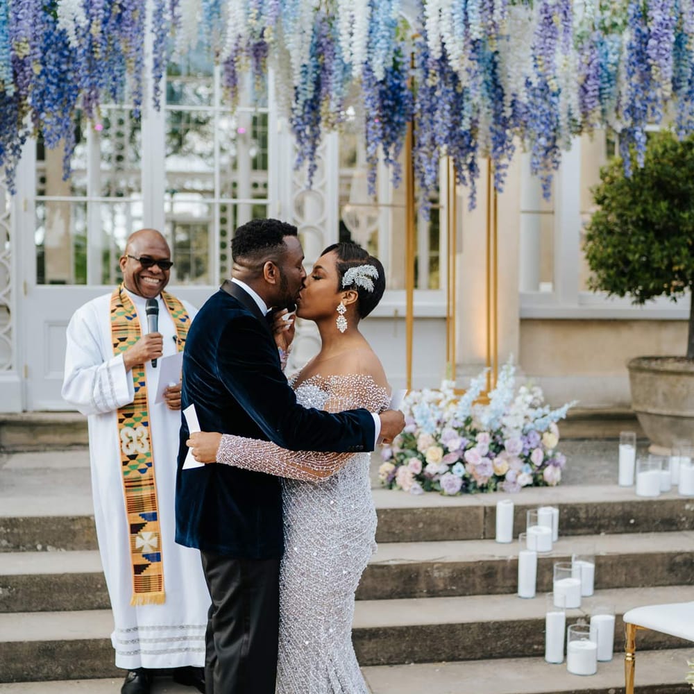 Groom in velvet midnight blue double breasted groom’s blazer, velvet bowtie and peach garden rose boutonniere kissing bride in illusion v neck sheer off shoulder long sleeve pearl embellished wedding gown standing underneath suspended pastel flower arrangements at Syon Park vow renewal 