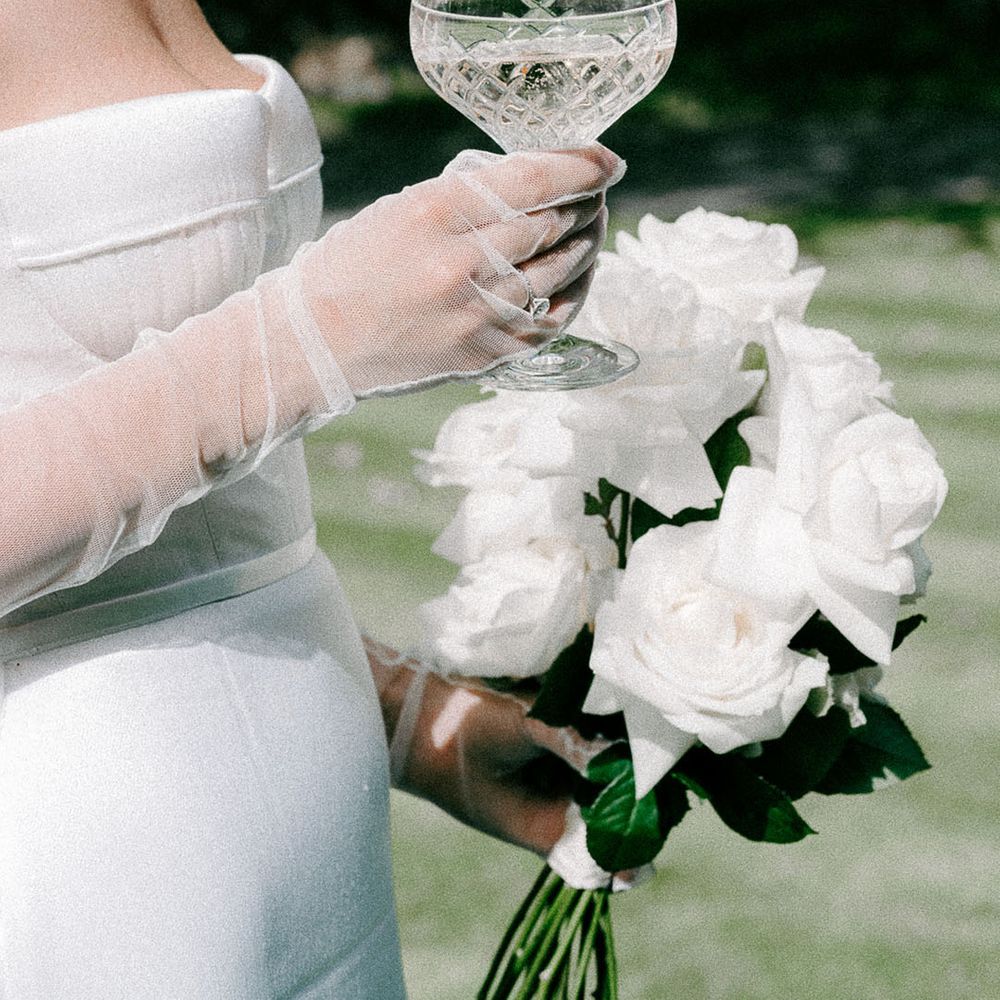 Bride with tulle sheer bridal gloves holding a champagne coupe and white rose long stem wedding bouquet 