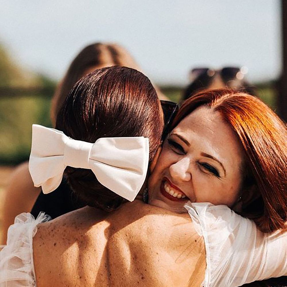 Image of bride hugging a guest while wearing a white bow hair accessory by Christophe Bourgeois Photography