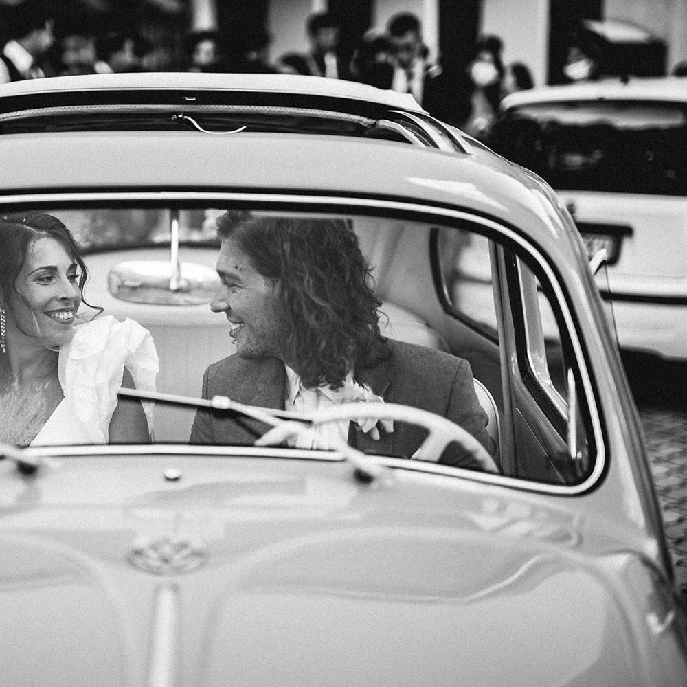 Bride with chandelier earrings and ruffle sleeves and groom with curly hair sitting in their wedding car 