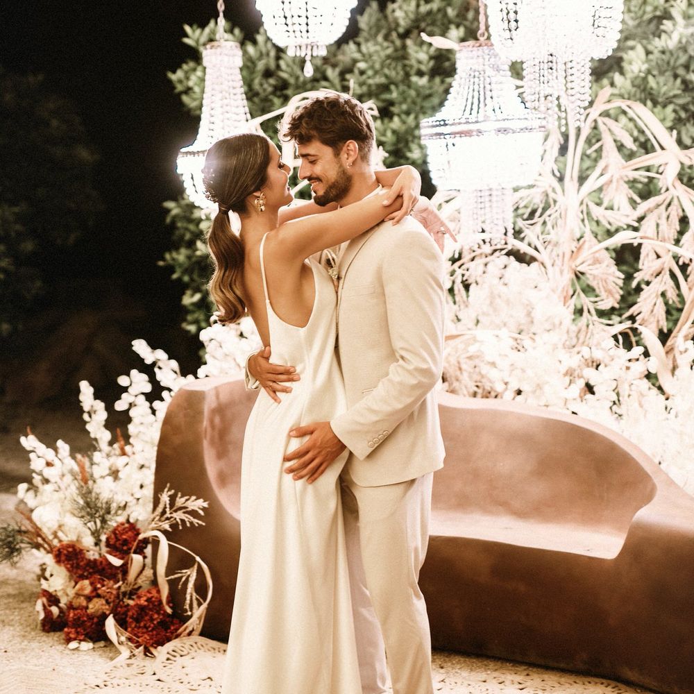 Bride in a satin slip dress kissing her groom in a beige suit under a canopy of chandelier lights 